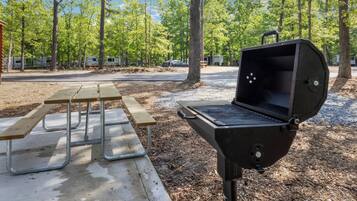 A barbecue grill and picnic table in a campground with trees and RVs in the background.