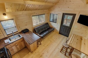 A compact kitchen with a sink, stove, and wooden cabinets. There is a black leather sofa and a small dining area with stools.