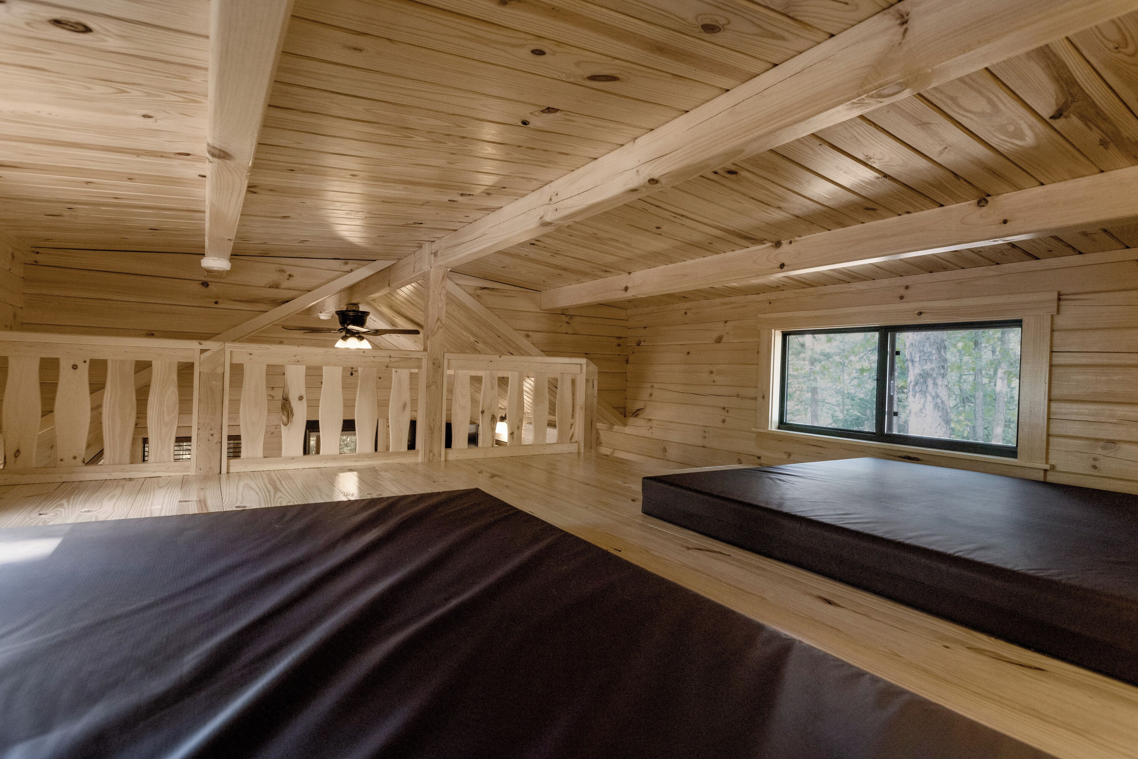 A cabin interior with a sloped ceiling, wooden beams, a window with a view of trees, and a bed with a dark mattress.