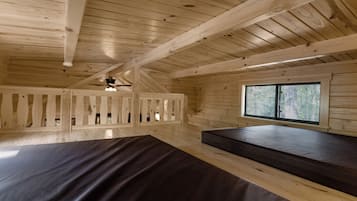 A cabin interior with a sloped ceiling, wooden beams, a window with a view of trees, and a bed with a dark mattress.
