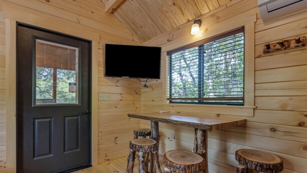 A wooden cabin interior with a dark door, a mounted flat-screen TV, and a window with blinds. - Sun Retreats Hospitality Creek (Monroe)