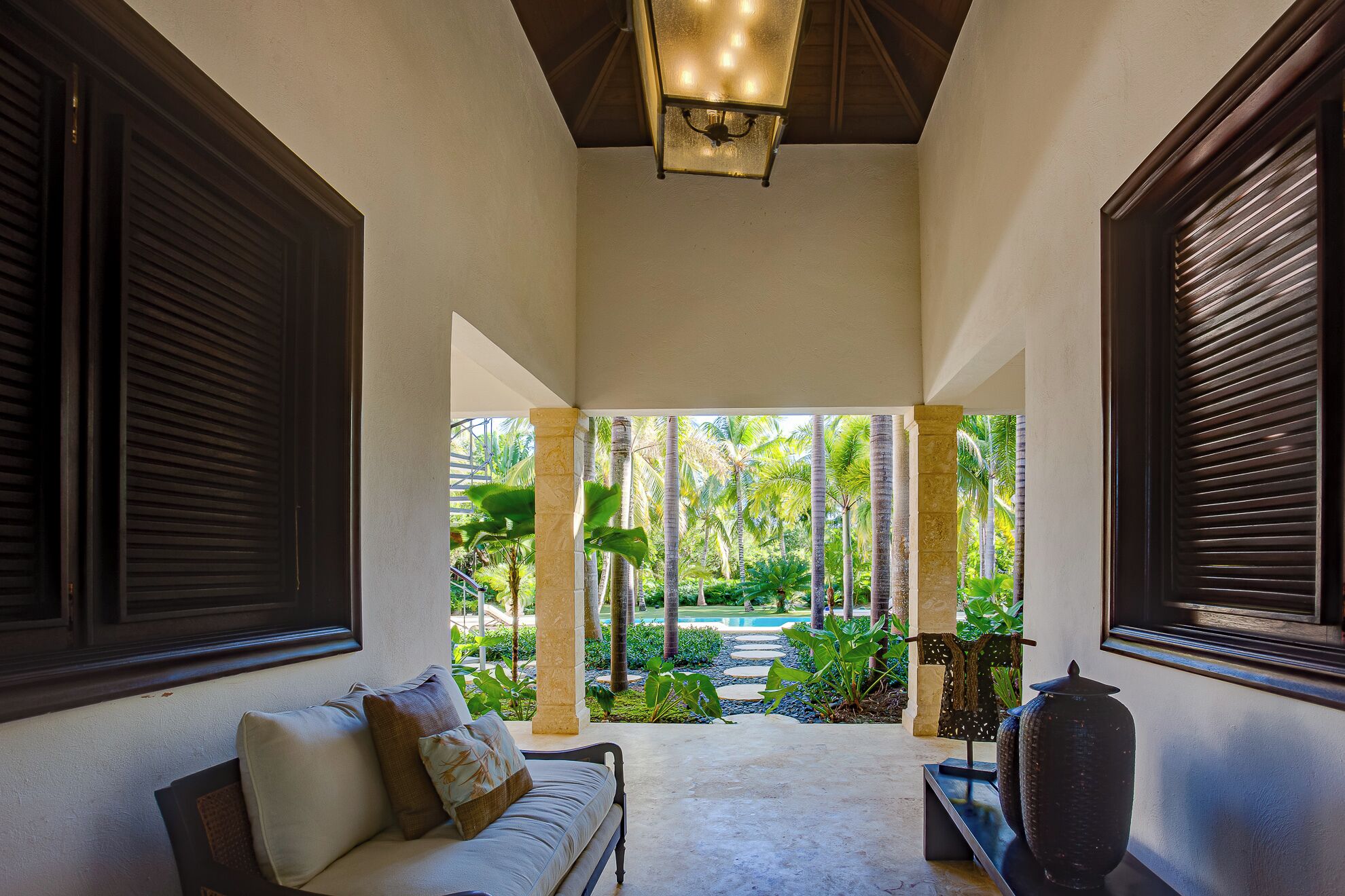 A covered outdoor seating area with a sofa, a television, and a view of a tropical garden.