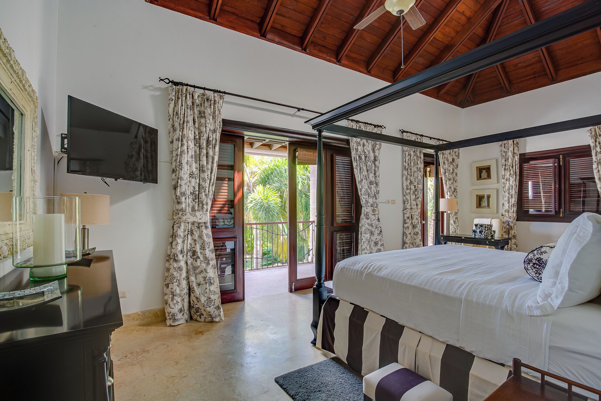 A bedroom with a bed, a television, a desk with a lamp, and a view of the outside.