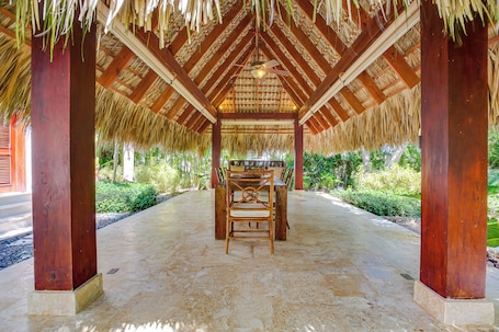 A covered outdoor seating area with a thatched roof, wooden pillars, and a concrete floor.