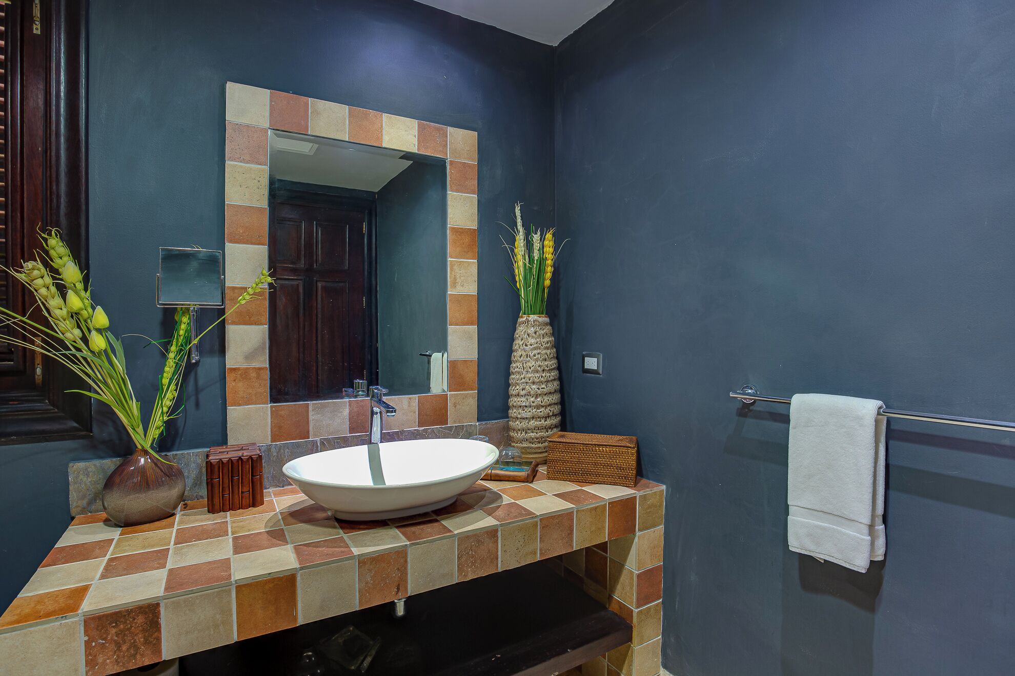 A bathroom with a tiled vanity, a large mirror, a towel rack, and a vase with plants.