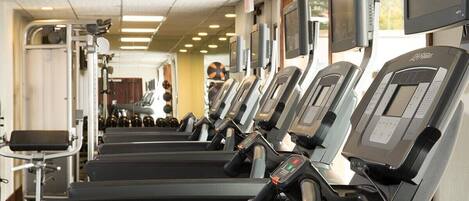 A row of treadmills in a gym with exercise equipment and a TV screen.
