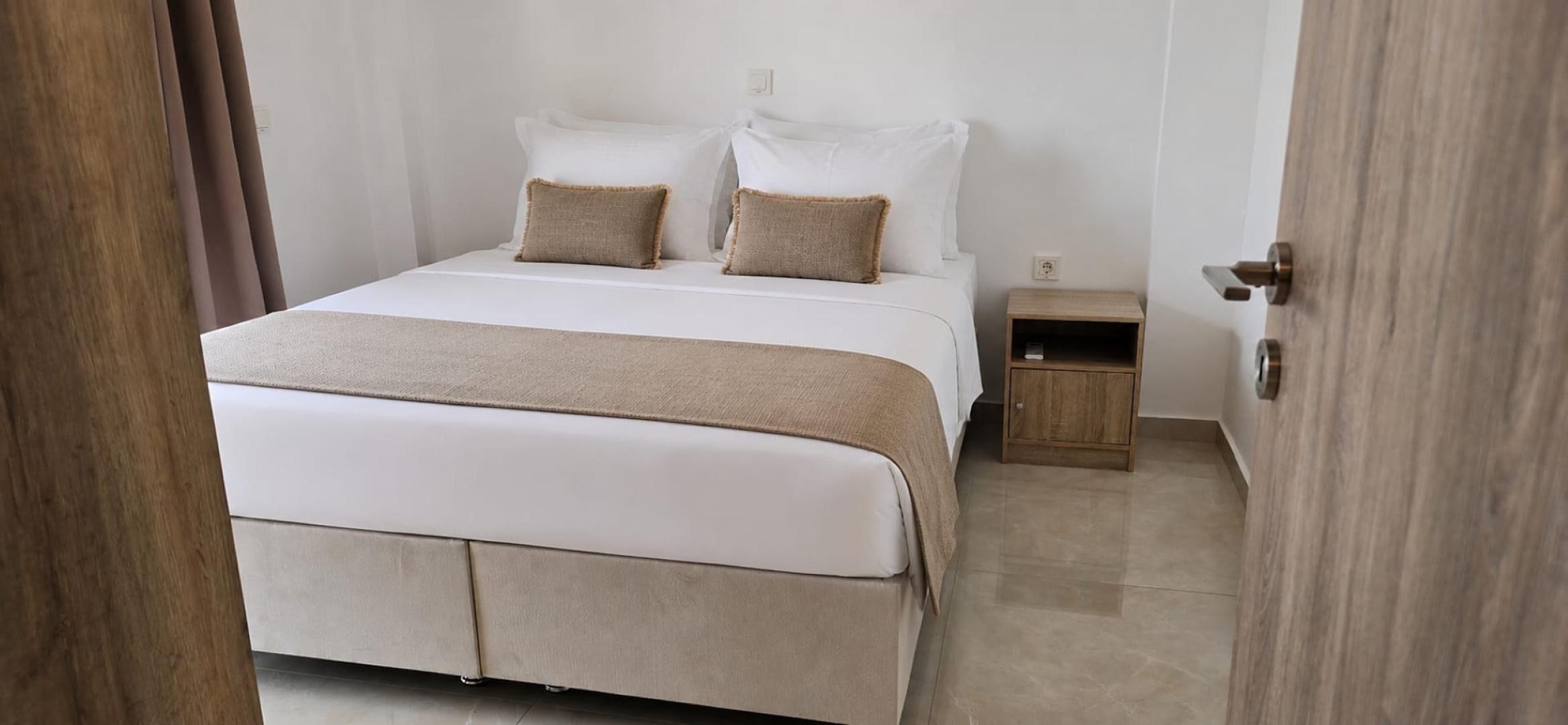 Een net opgemaakt bed met een beige deken en kussens, een houten nachtkastje met een lamp, en een deur die naar een andere kamer leidt.