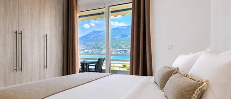 A hotel room with a bed, a view of the sea and mountains, and a balcony with a table and chairs.