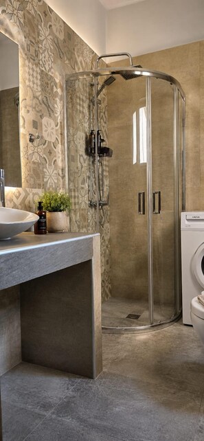 A modern bathroom with a glass-enclosed shower, a sink with a basin, a potted plant, and a washing machine. - Esperides Pearl Luxury SeaView Apartment by DM Hospitality (LOUTRAKI)