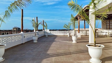 A rooftop terrace with potted palm trees, a white railing, and a view of the ocean.