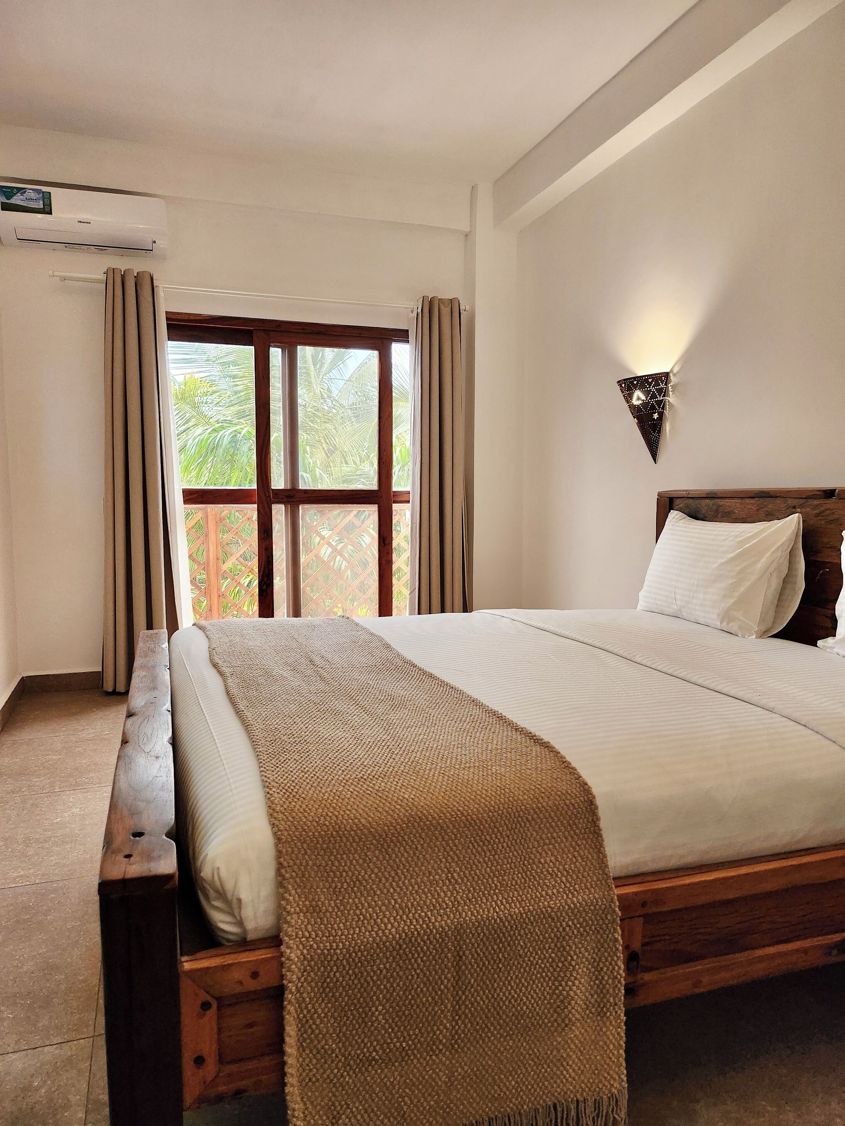 A bedroom with a wooden bed, a window with curtains, a wall-mounted light fixture, and an air conditioning unit.