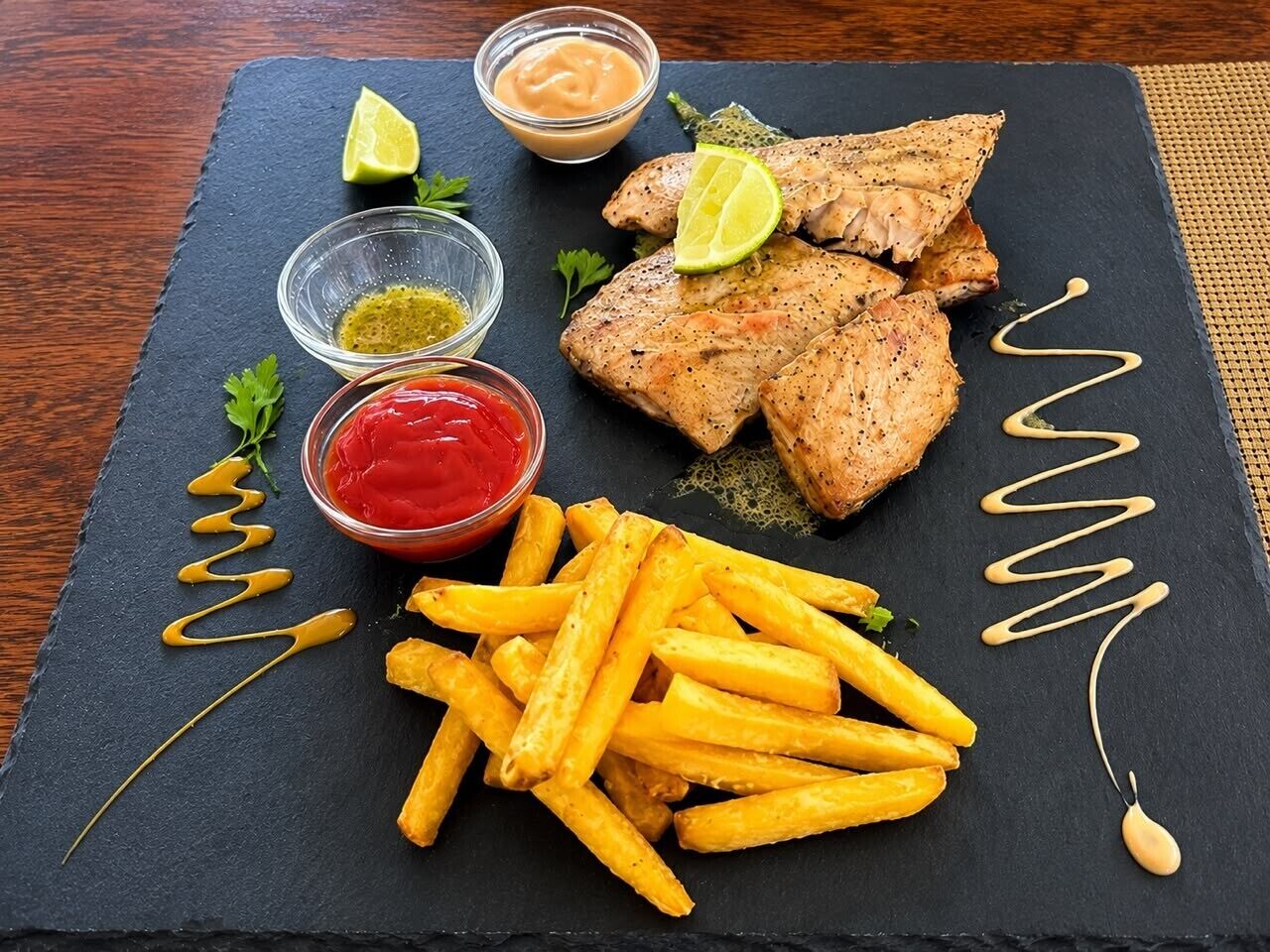 Un plato de pescado a la parrilla y papas fritas sobre una tabla de pizarra con salsas.