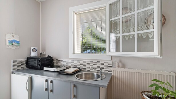 A compact kitchen with a microwave, toaster, and sink. There's a window with decorative lace curtains and a view of greenery outside.