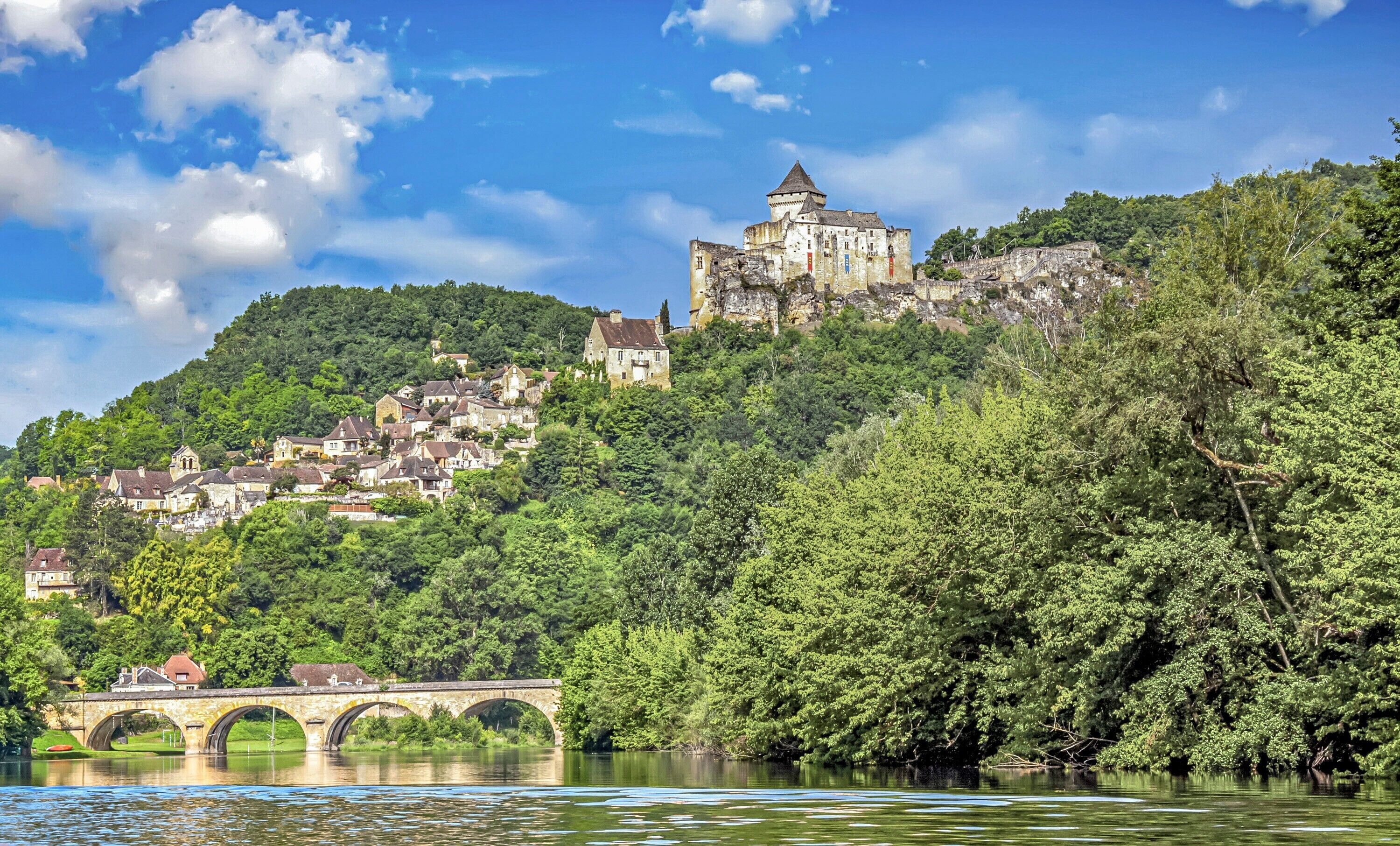 Un château historique, perché sur une colline qui surplombe une rivière, avec un pont en premier plan.