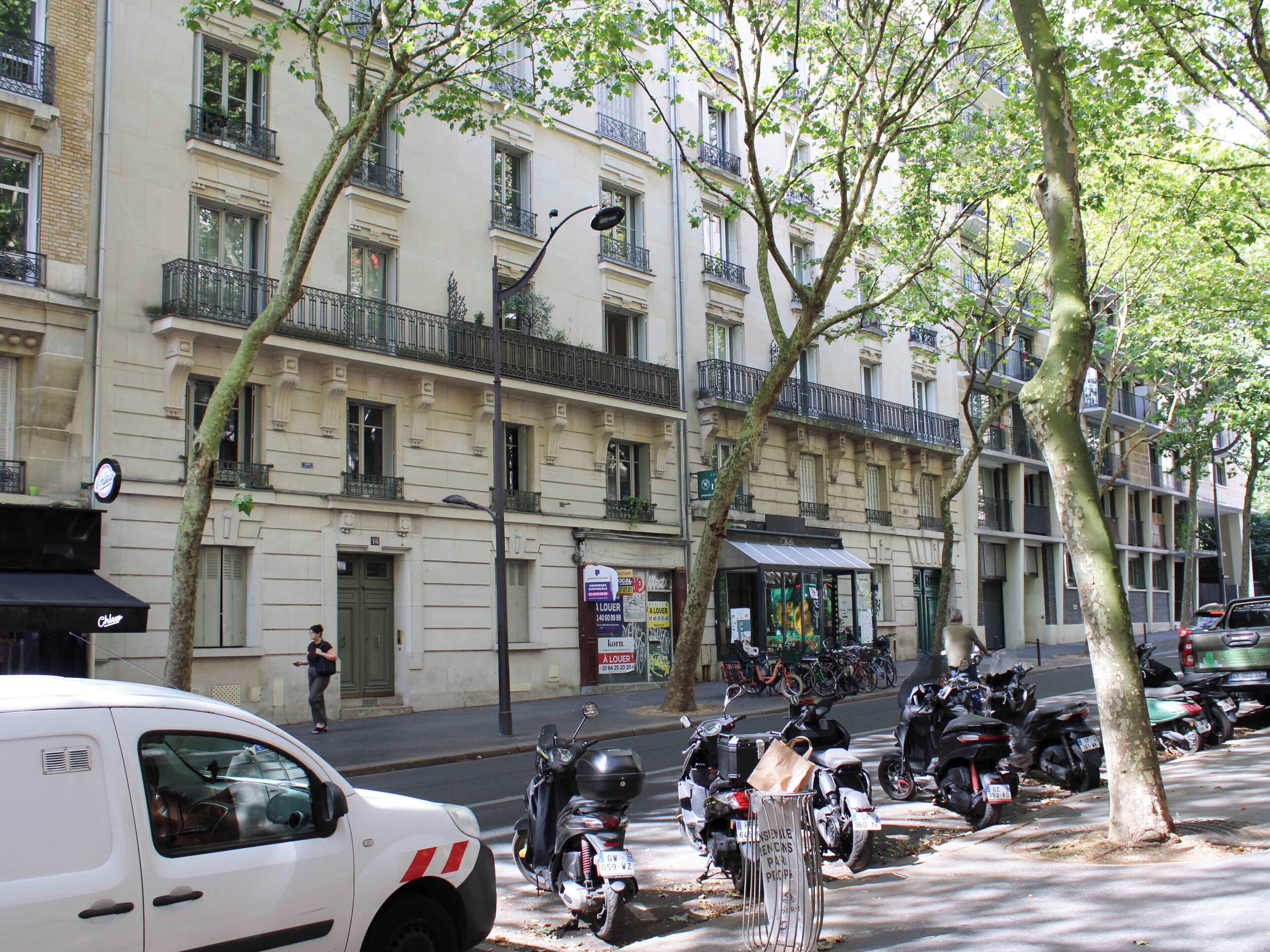 A street with parked scooters, a white car, and a building with multiple balconies and windows.