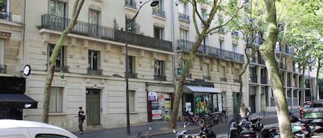 A street with parked scooters, a white car, and a building with multiple balconies and windows.