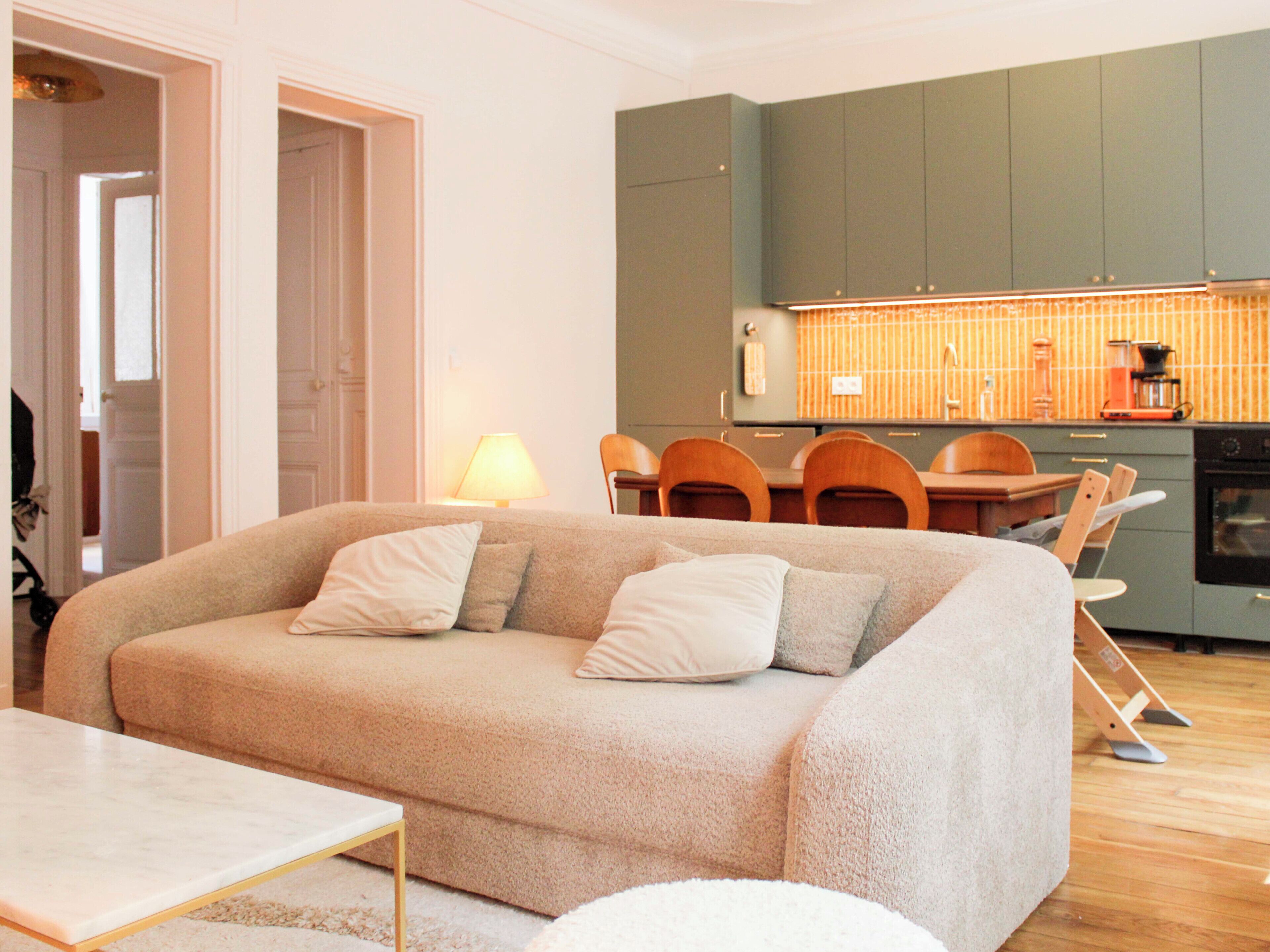 A living room with a sofa, a dining table, and chairs. There is a kitchen area with cabinets and a stove.