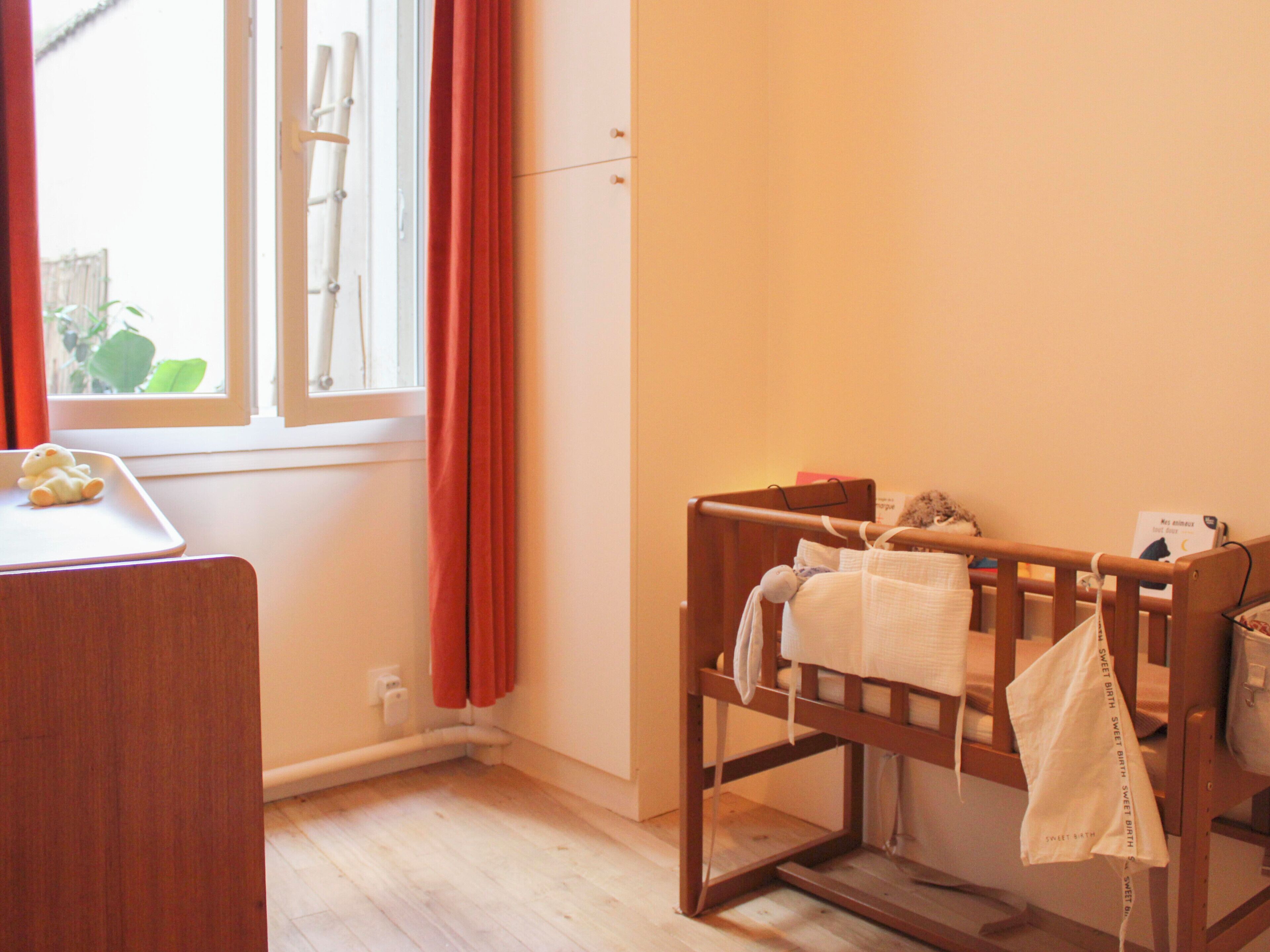 A room with a wooden crib, a small table with a teddy bear, and a window with curtains.