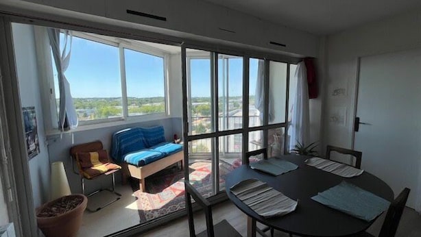 Quarto com uma grande porta de vidro deslizante que dá para uma varanda, uma mesa de jantar redonda com cadeiras, um sofá e um vaso de planta.