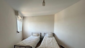 Acomodação pequena, com um quarto e duas camas de solteiro, janela com cortinas e luminária.