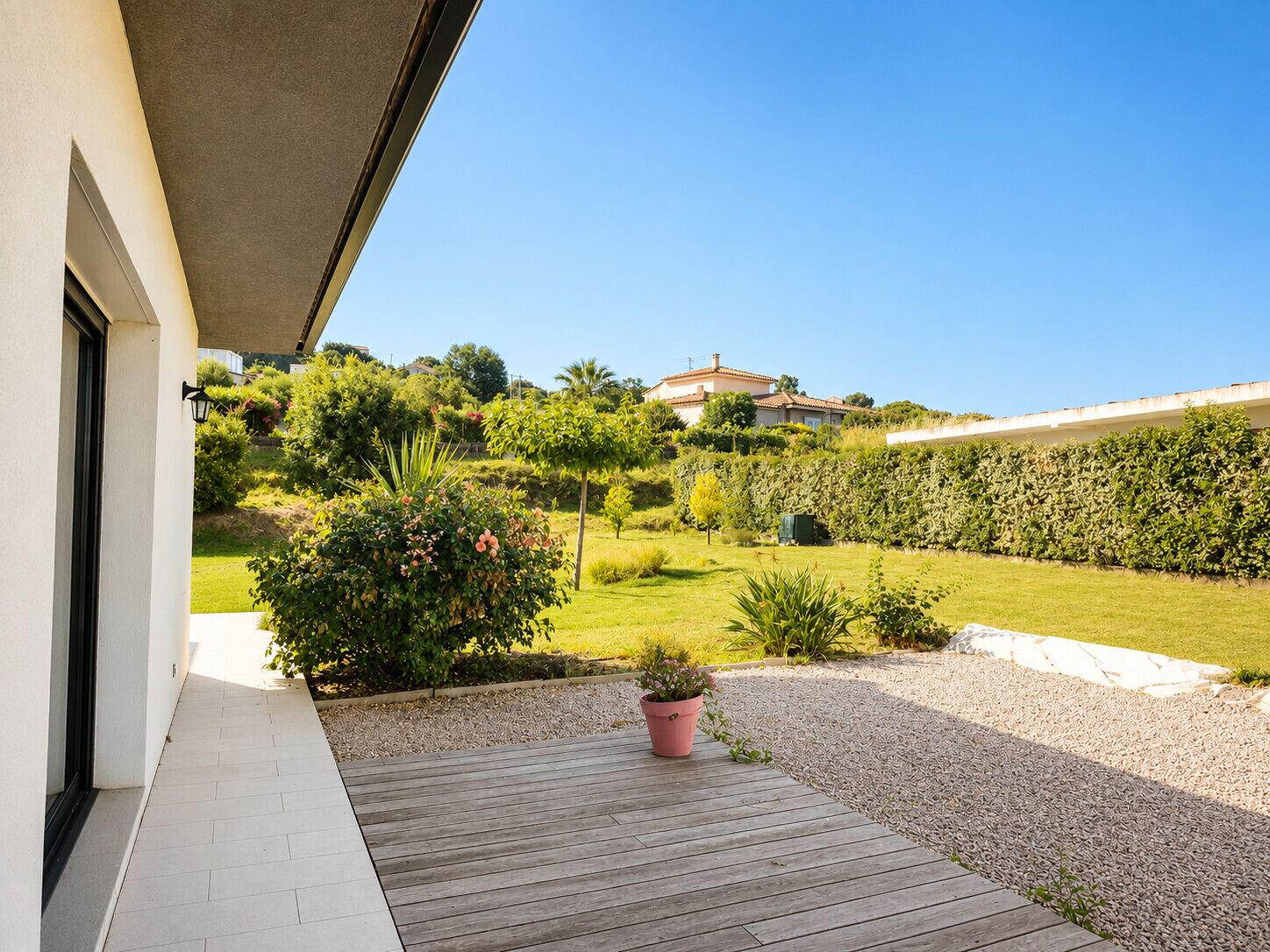Un bâtiment moderne doté d’une terrasse en bois, d’un jardin avec différentes espèces de plantes et d’un ciel d’un bleu éclatant.