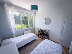 A bedroom with two beds, a window with curtains, a bookshelf, and a door.