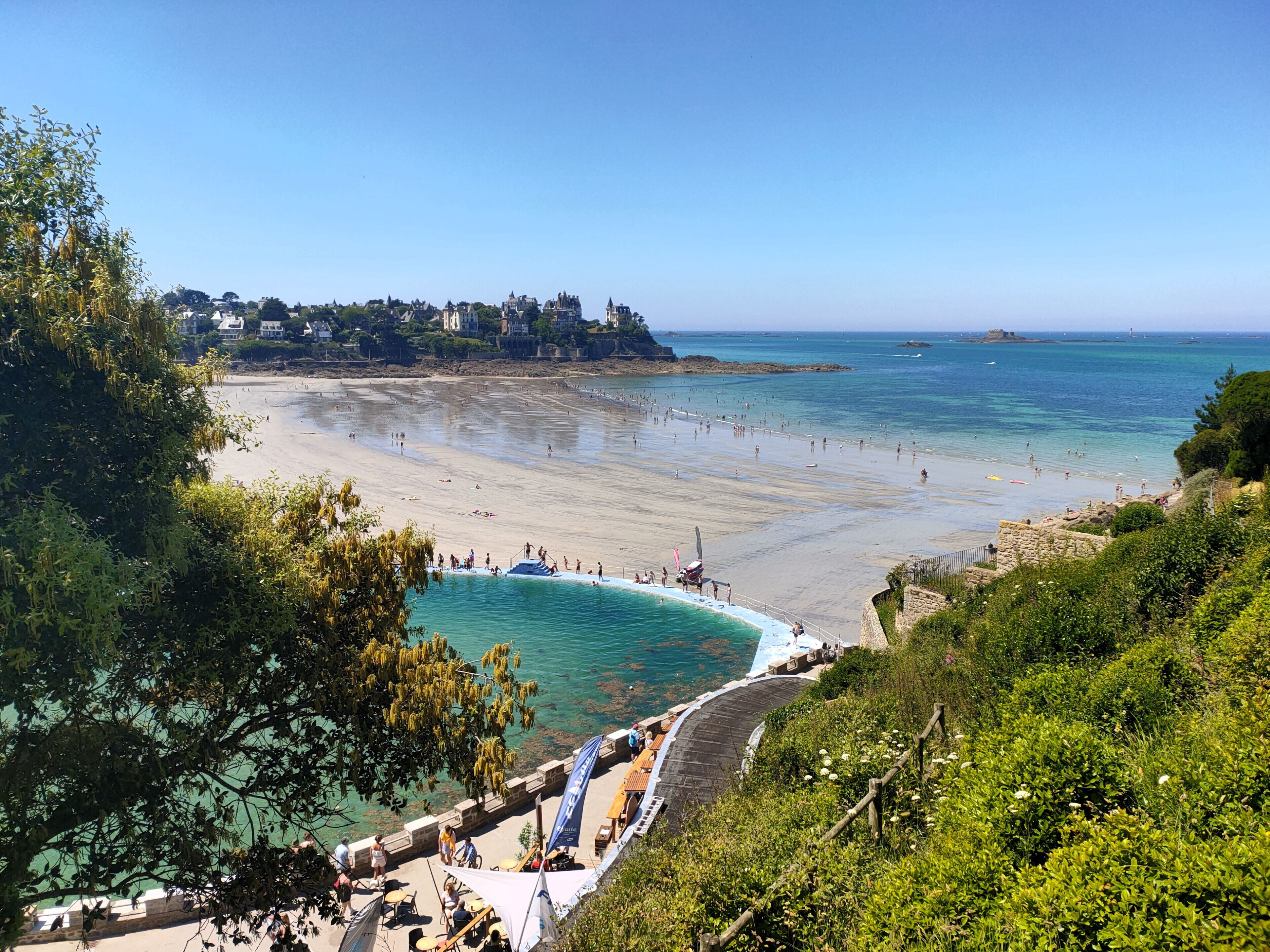 A coastal beach with a clear view of the sea, a swimming pool, and a stone pathway leading to the beach.