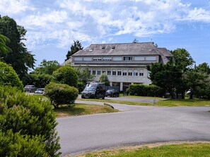 Ein großes Gebäude mit zahlreichen Fenstern und einem geneigten Dach, umgeben von Grünflächen und parkenden Autos.