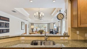 A kitchen with granite countertops, a stainless steel sink, wooden cabinets, a chandelier, and a wall-mounted clock. - New! Epic Reunion Family Retreat (Kissimmee)