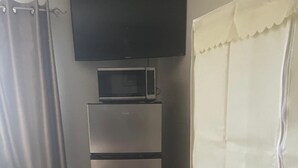 A compact room with a flat-screen TV mounted above a stainless steel refrigerator, a white chair, and a white curtain.