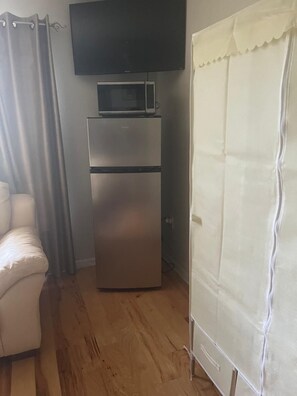 A compact room with a flat-screen TV mounted above a stainless steel refrigerator, a white chair, and a white curtain. - Clean & Cozy Room with Private Bath. Minutes to Disney. Free parking. (Kissimmee)