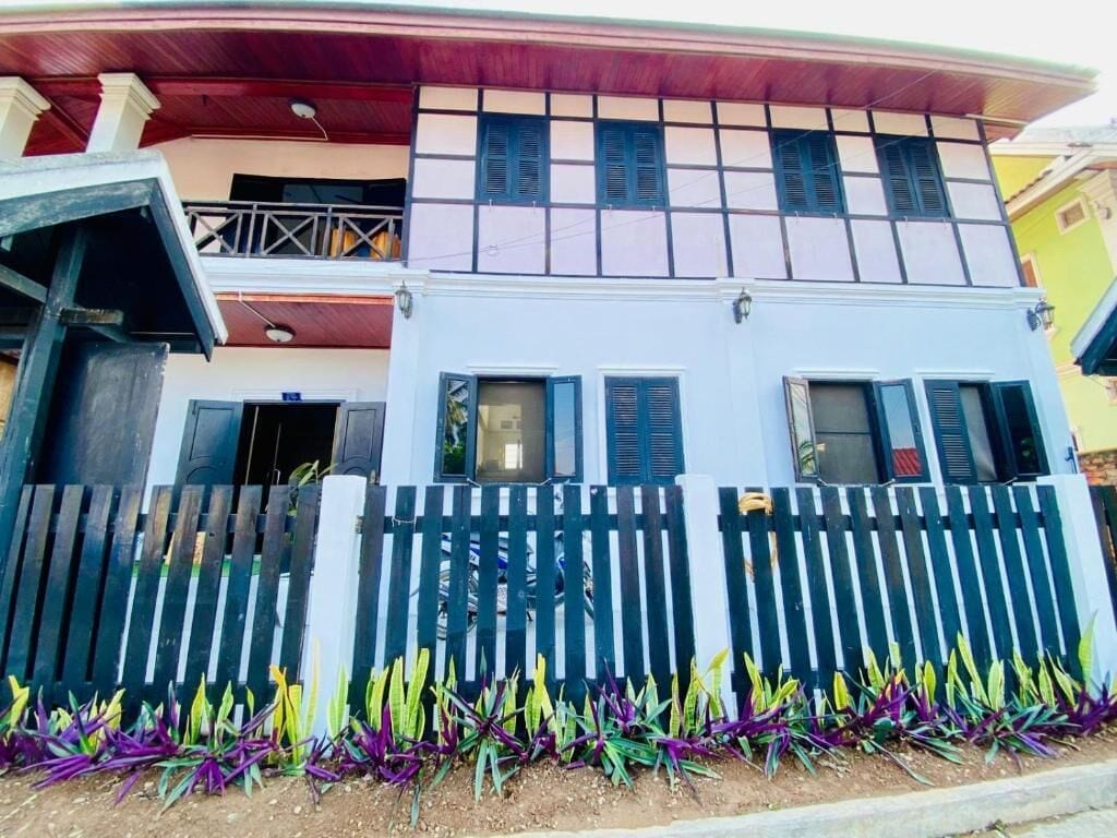 A two-story building with a balcony on the upper floor, a wooden fence in front, and a variety of plants at the base.