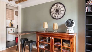 Ein Zimmer mit einem Holztisch, zwei schwarzen Hocker, einem Holzschrank mit Glastüren, einer Wanduhr, einem Ventilator und einem Bücherregal.