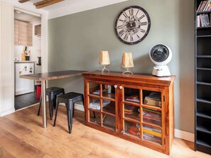 Ein Zimmer mit einem Holztisch, zwei schwarzen Hocker, einem Holzschrank mit Glastüren, einer Wanduhr, einem Ventilator und einem Bücherregal.