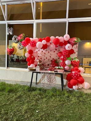 Une table décorée de ballons et de fraises, installée à l’extérieur d’un bâtiment doté de grandes fenêtres.