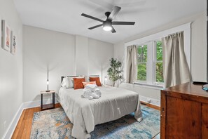 A bedroom with a bed, a ceiling fan, a window with curtains, a bedside table with a lamp, and a wooden dresser. - Charming Garden Home in Old Louisville (Louisville)