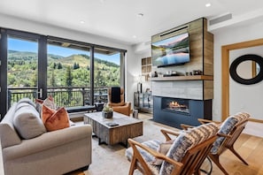 A modern living room with a fireplace, large TV, and a balcony with a scenic view. - Riverfront Lodge Condo W/Resort Amenities (Avon)