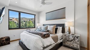 Ein modernes Schlafzimmer mit einem großen Bett, einem Nachttisch, einem Deckenventilator und einem Fenster mit Blick auf Gebäude und Bäume.