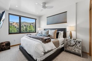 Ein modernes Schlafzimmer mit einem großen Bett, einem Nachttisch, einem Deckenventilator und einem Fenster mit Blick auf Gebäude und Bäume.