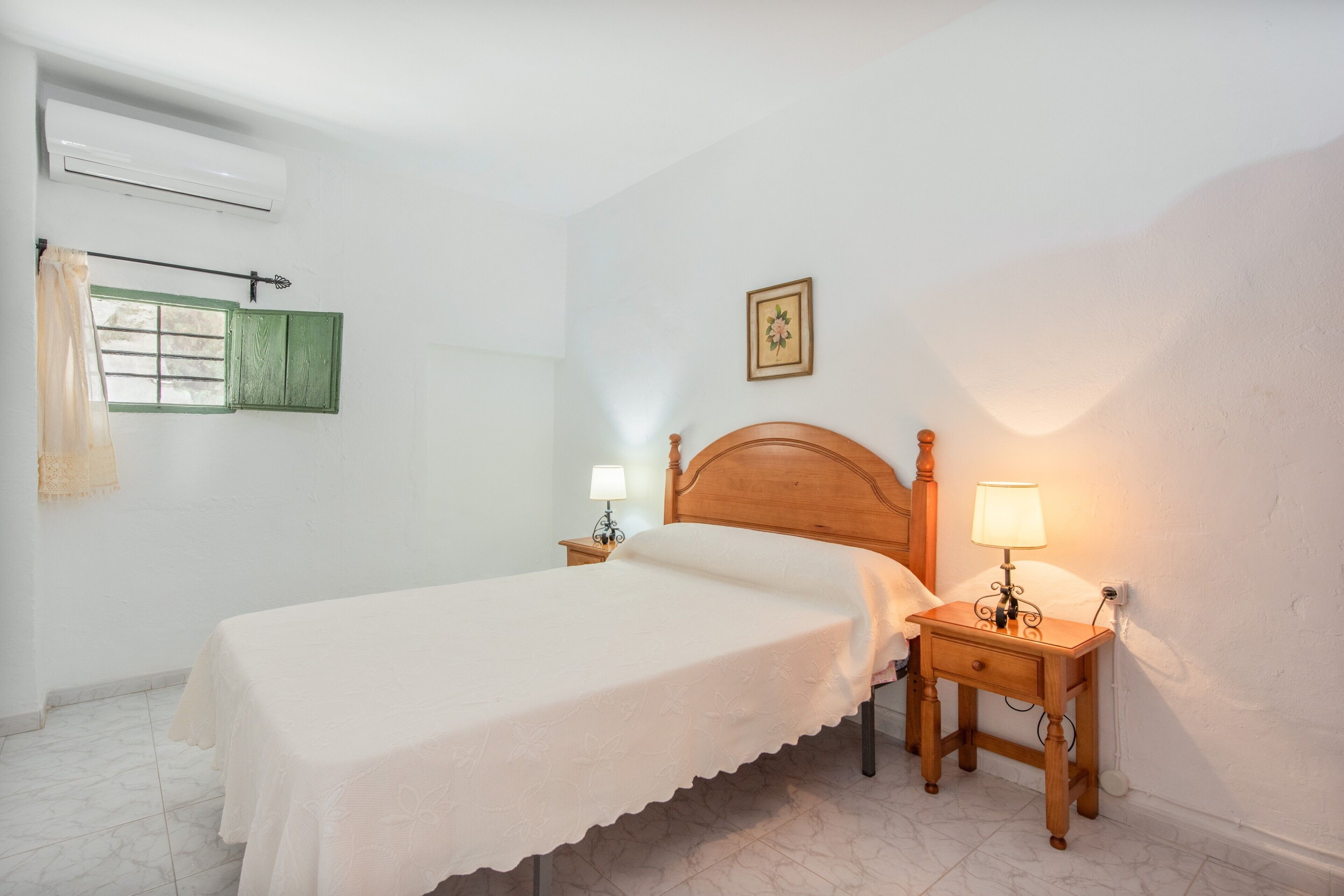 Quarto com uma cama de solteiro, duas mesas de cabeceira com luminárias, uma janela com persianas e um ar-condicionado fixado na parede.