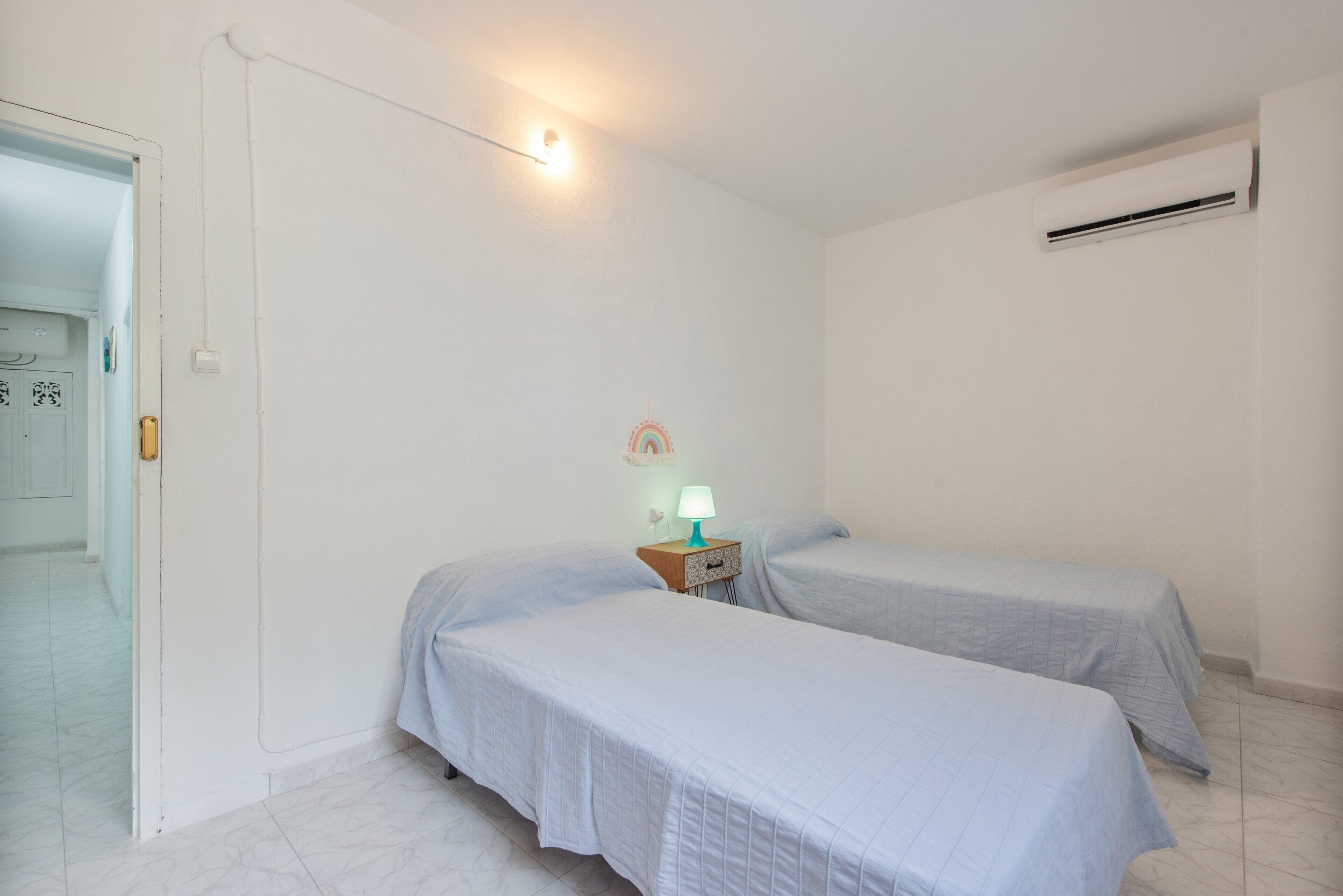 Una habitación sencilla con dos camas individuales, una mesita de noche con lámpara, un equipo de aire acondicionado y una puerta con pomo.