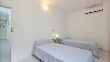 Una habitación sencilla con dos camas individuales, una mesita de noche con lámpara, un equipo de aire acondicionado y una puerta con pomo.