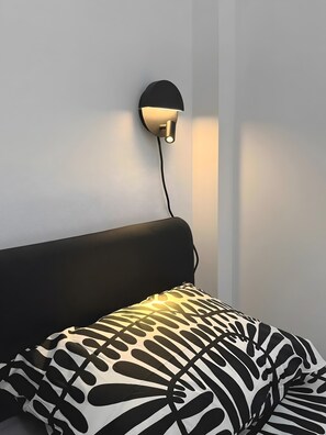 Ein modernes Bett mit schwarzem Kopfteil und einem gemusterten Kopfkissen, einer an der Wand montierten Leuchte und einem sichtbaren Stromkabel.