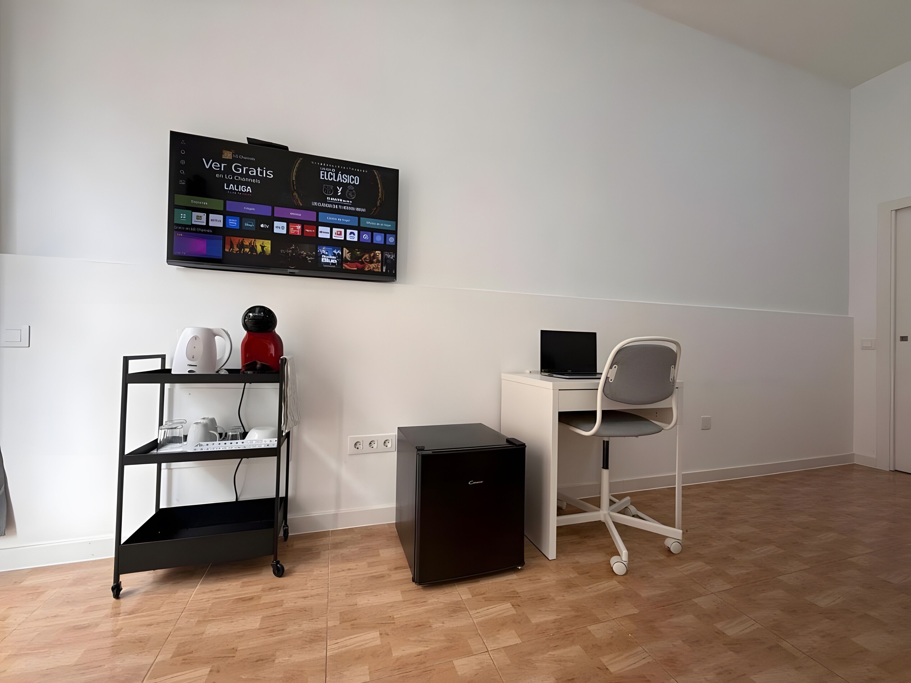 Una stanza con una scrivania, una sedia, un altoparlante nero, un piccolo mobiletto nero, uno scaffale bianco con oggetti vari e una TV a parete che mostra diverse applicazioni.