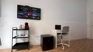 Una stanza con una scrivania, una sedia, un altoparlante nero, un piccolo mobiletto nero, uno scaffale bianco con oggetti vari e una TV a parete che mostra diverse applicazioni.