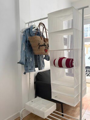 Ein Zimmer mit einem weißen Kleiderschrank, einer Kleiderstange mit Jeans und einer gewebten Tasche, einer weißen Aufbewahrungseinheit mit Regalen und einem schwarzen Sofa.
