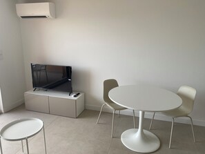 Ein modernes Zimmer mit einem Flachbildfernseher auf einer weißen Kommode, einem runden Tisch mit zwei Stühlen und einer Klimaanlage an der Wand.