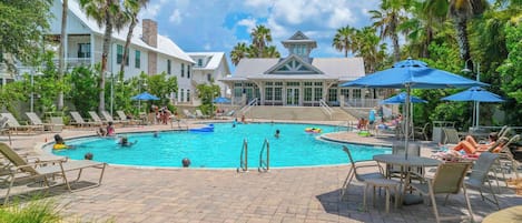 A pool area with lounge chairs, umbrellas, and a building with a white facade in the background.