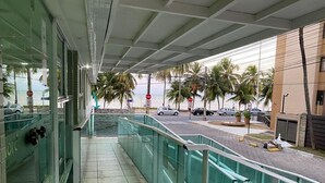 A covered walkway with glass railings overlooking a street with palm trees and parked cars. - Located across from Cabo Branco Beach - Apartment with ocean view. (João Pessoa)