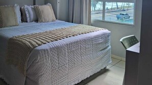 A hotel room with a bed, a window with a view of palm trees, and a chair. - Located across from Cabo Branco Beach - Apartment with ocean view. (João Pessoa)